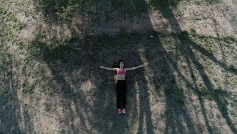 Ascending-Drone-Shot-of-Young-Brunette-Woman-lying-on-the-ground-with-arms-open-next-to-a-tree