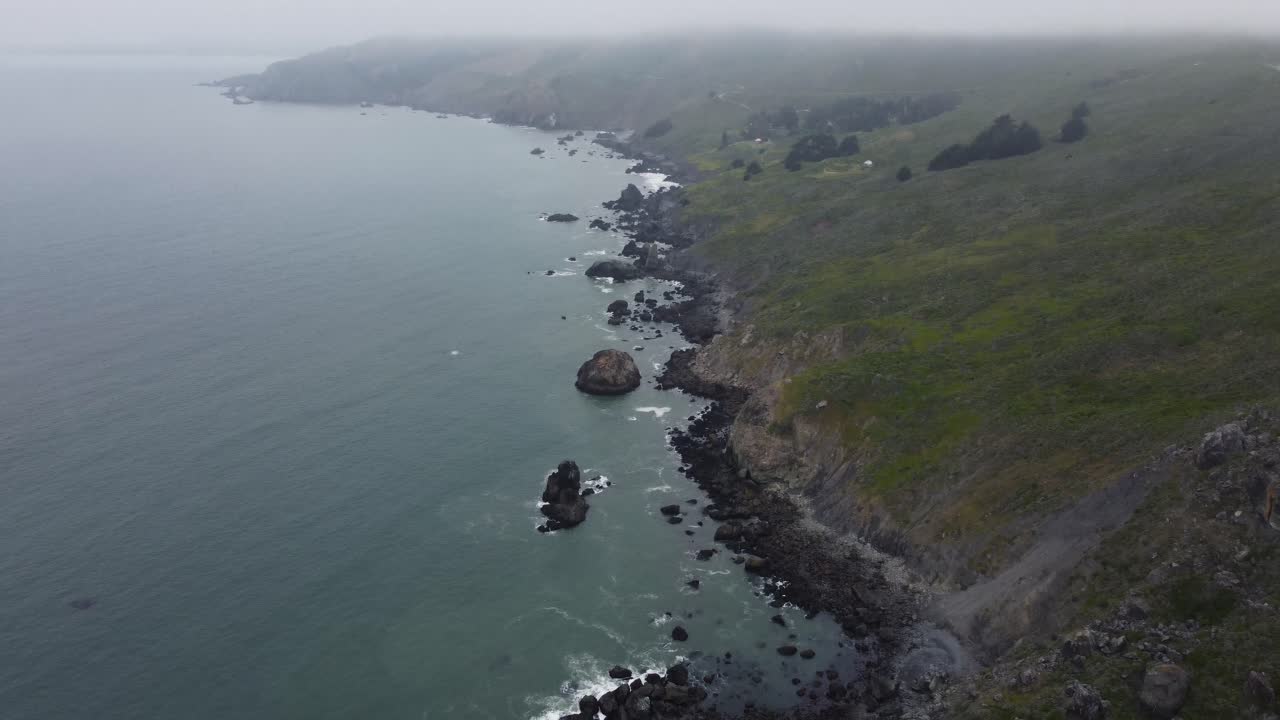 Slow reveal shot of the rocky California coastline on a foggy day in Summer. The shot shows an aerial view of the waves crashing on the rocks before panning to reveal the green, shrub covered cliffs.