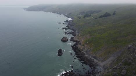 slow reveal shot of the rocky california coastline on a foggy day in summer. the shot shows an aerial view of the waves crashing on the rocks before panning to reveal the green, shrub covered cliffs.