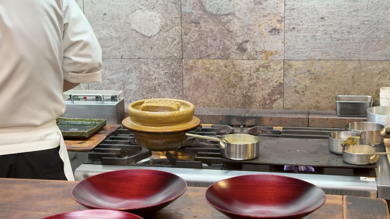Một đầu bếp chuyên nghiệp mặc đồng phục trắng đang chuẩn bị thức ăn bằng nồi đất nung donabe truyền thống.