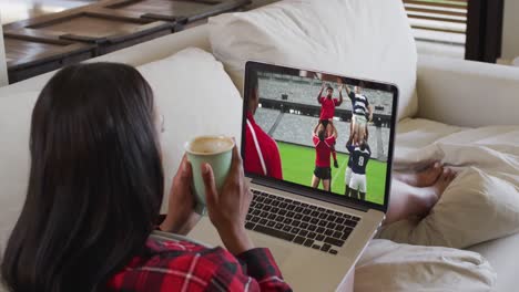 compuesto de una mujer sentada en casa sosteniendo un café viendo un partido de rugby en una computadora portátil