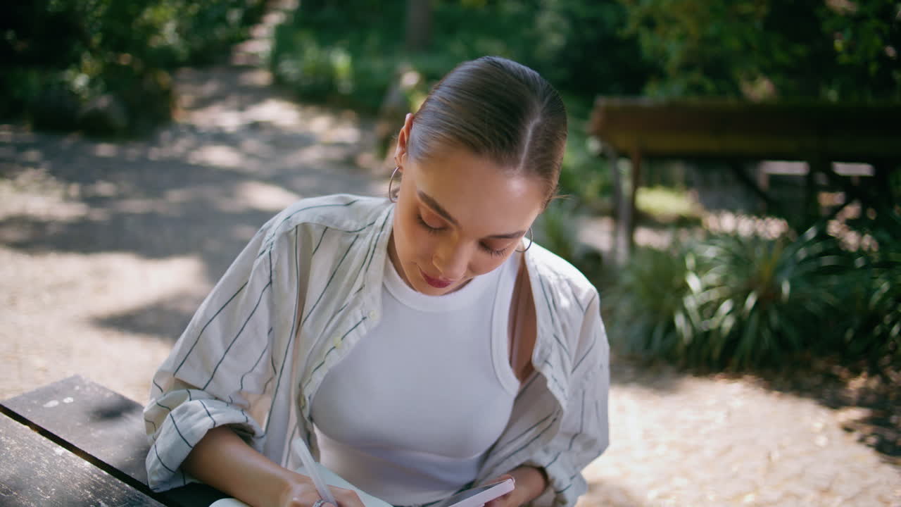 hermosa mujer creando texto en el bloc de notas sentada en el parque con el teléfono inteligente de cerca.