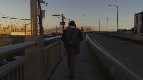 Following a travelling man walking along the sidewalk towards a bridge in the city while cars pass by in the evening