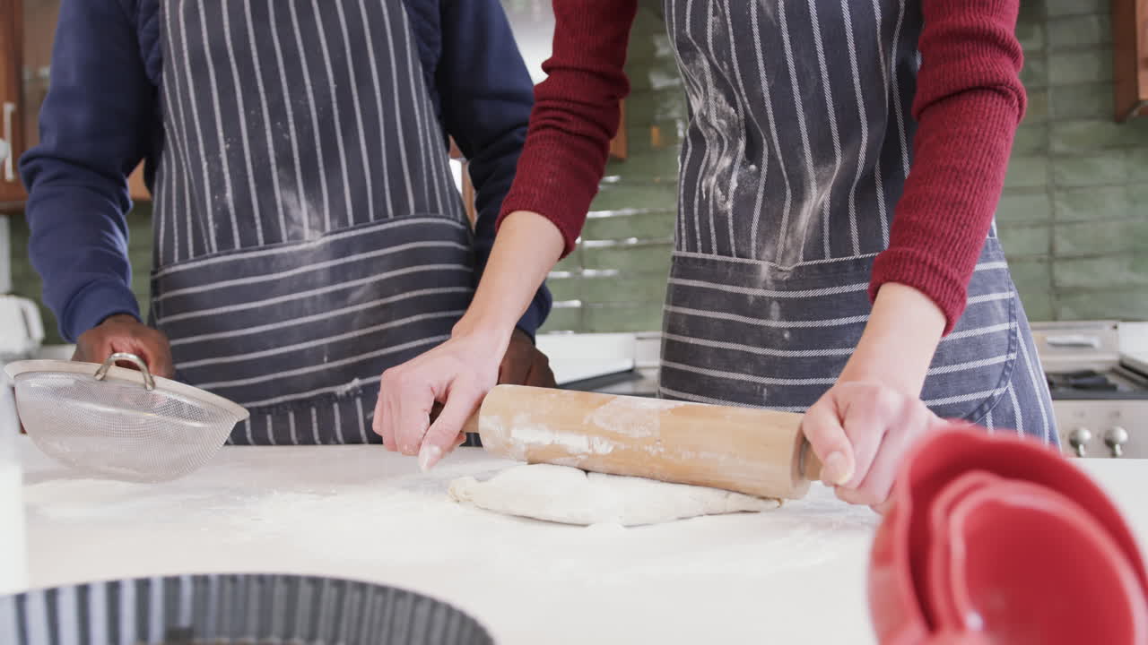 Happy diverse couple standing in kitchen, sifting flour and rolling dough,slow motion