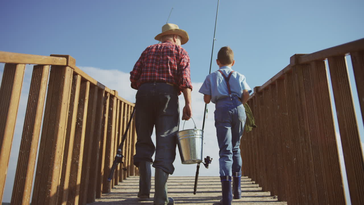 Rear view of a senior man and his small grandson walking on the wooden bridge with equipment for fishing