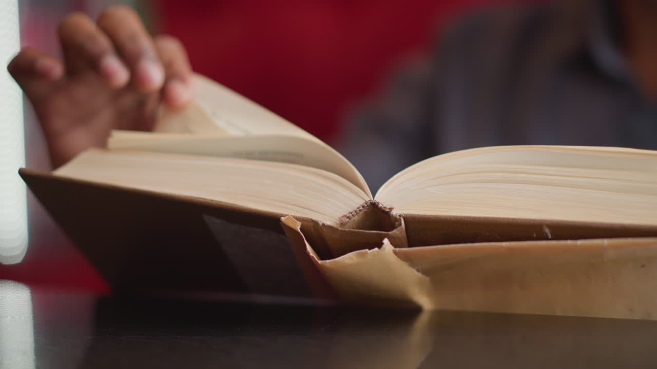 Close-up of male turning page of thick open book on dark surface, with focus on torn book jacket and fingers in motion, suggesting moment of reading, learning, or relaxed quiet study in casual indoor setting