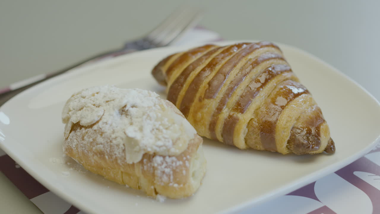 Close-up of two pieces of fresh pastry on a white plate: one glazed croissant and another covered with icing sugar