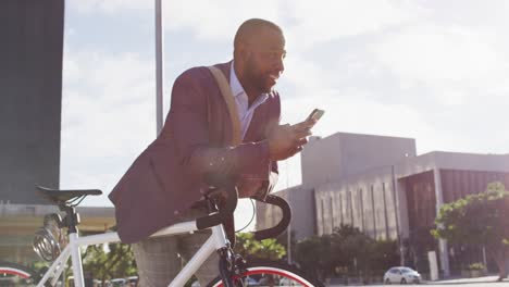 Hombre-Afroamericano-En-La-Ciudad,-Sentado-En-Bicicleta-Bajo-El-Sol-Usando-Un-Teléfono-Inteligente