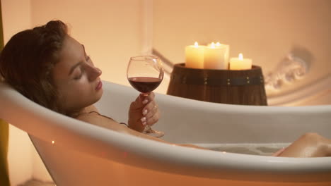 woman bathing with pleasure lying down in the tub with foam and drinking red wine spending time in luxury spa resort