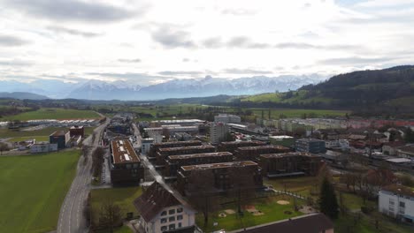 Ciudad-Suiza-Con-Casas-Pintorescas-Con-Un-Telón-De-Fondo-De-Majestuosos-Alpes-Nevados,-Cielos-Nublados,-Vista-Aérea