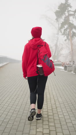 back view of woman in red winter jacket and boots carrying backpack and headphones around neck, walking along snow-dusted path lined with benches and trees in calm foggy winter atmosphere