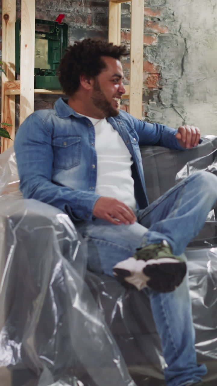 Happy African-American guys discuss room restoration ideas sitting on sofa covered with plastic sheet. Black friends prepare to repair house