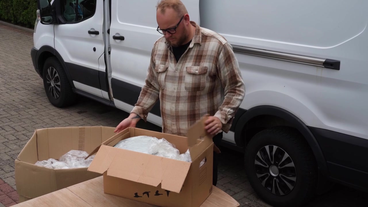 Muž stojí vedle bílé dodávky a třídí staré plastové výplně do kartonových krabic pro opětovné použití v ekologicky šetrné balíkové přepravě.