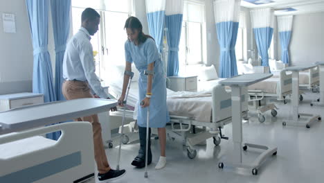 diverse male doctor helping happy female patient walk with crutches in ward, copy space, slow motion