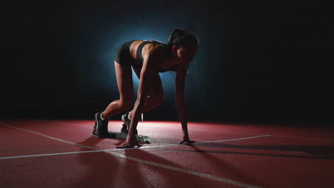 slender young girl athlete is in position to start running in the pads on the track in slow motion