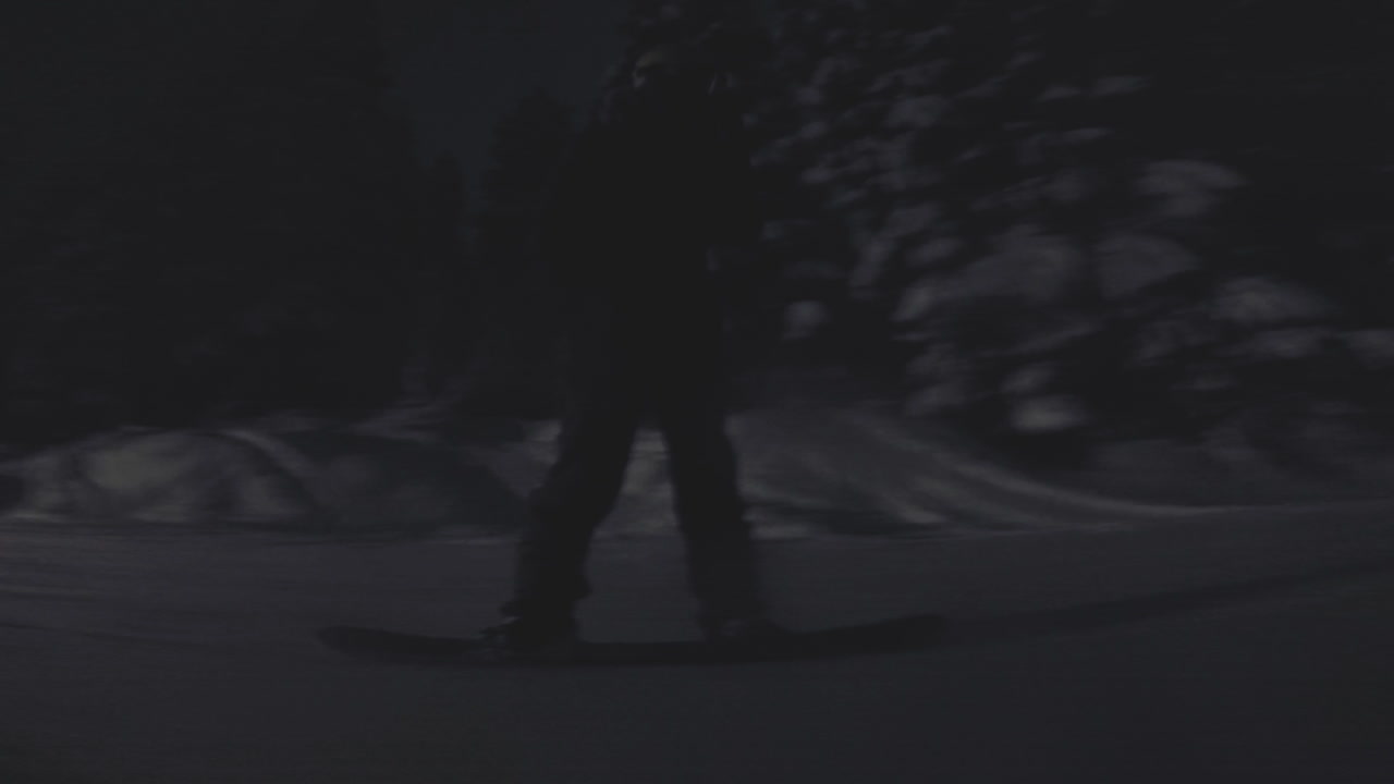 Silhouetted man snowboards down a mountain at night. Lights shine on the snowy park.