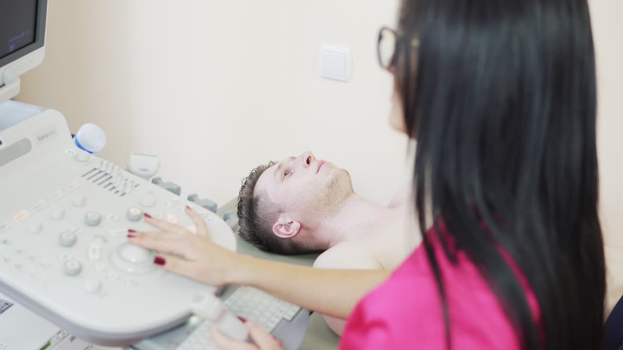 Close up view of young female doctor with long hair in rose uniform is screaning her patient's heart with ultrasonography, looking at the screen, and consulting.