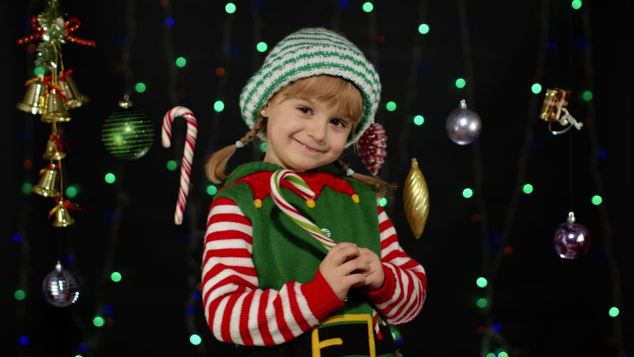 Malá dívka v kostýmu vánočního skřítka, pomocníka Santa Clause, líže lízátko ve tvaru cukrové hole a karamelové sladkosti