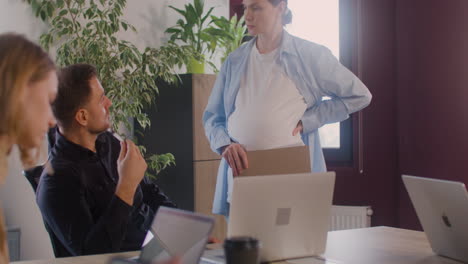 pregnant woman is explaining a project talking with a coworker in a meeting room while she is touching her belly 3