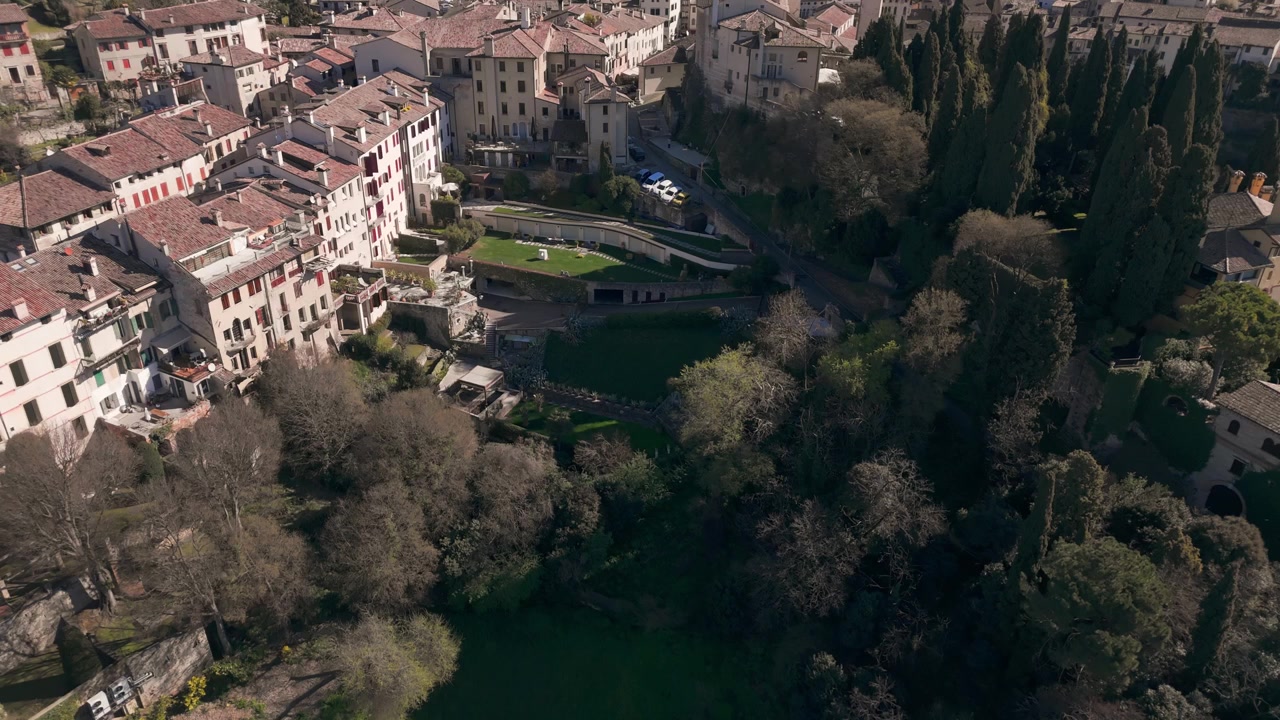 Vista aérea flutuando sobre edifícios históricos e árvores de Asolo em um dia claro.