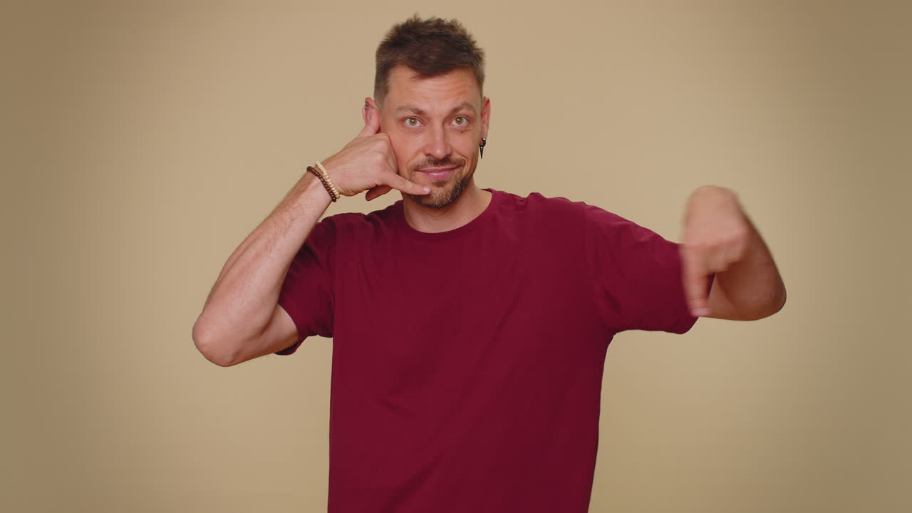 수염이 있는 잘생긴 젊은이가 카메라를 바라보며 전화 제스처를 하고 있다.