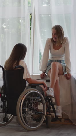 blonde woman sits on windowsill talking to girl partner with disability in wheelchair after surgery. lesbian couple enjoys nice conversation at home