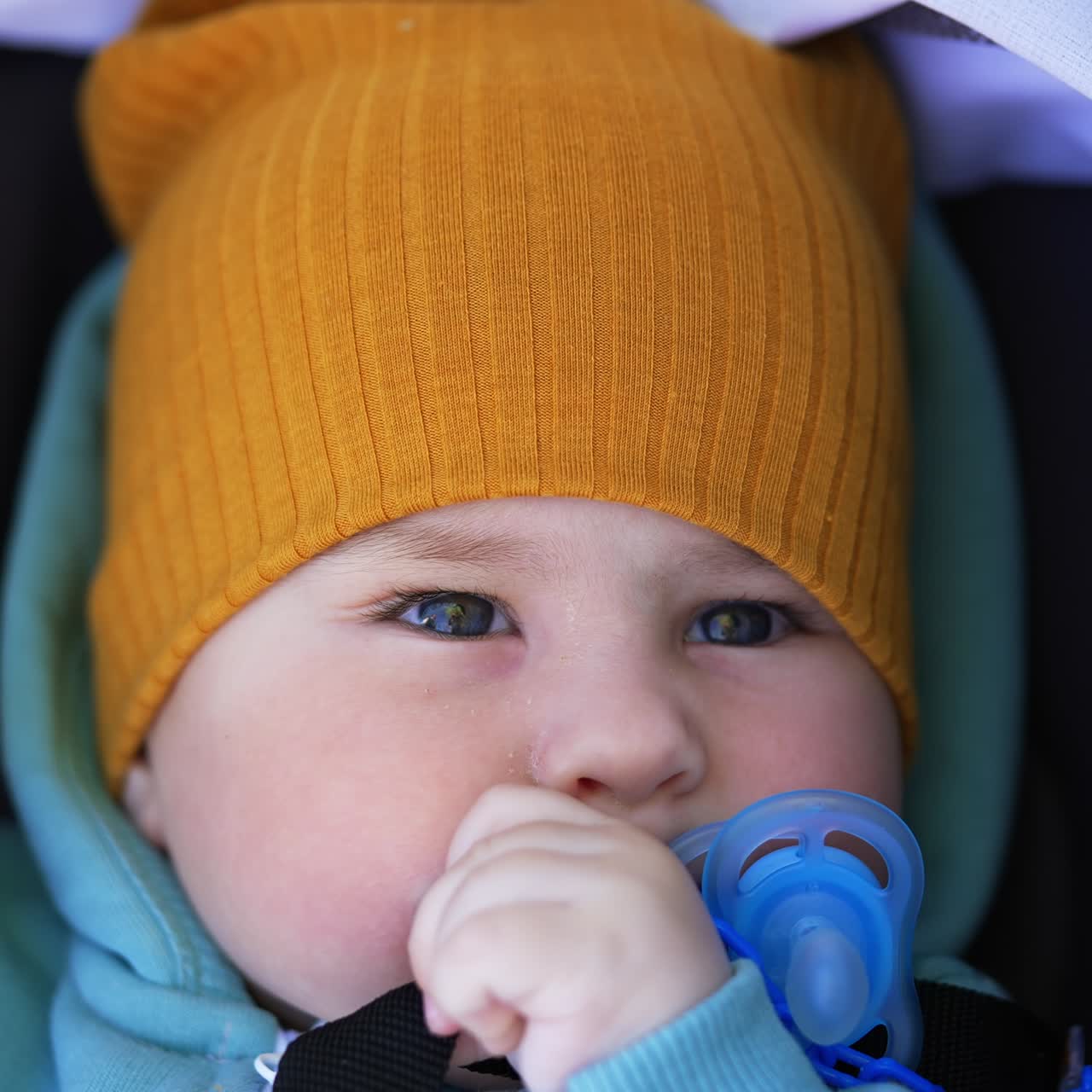 Grey-eyed toddler wearing yellow cap blinking eyes slowly. Lovely boy holding pacifier at his mouth lying in stroller