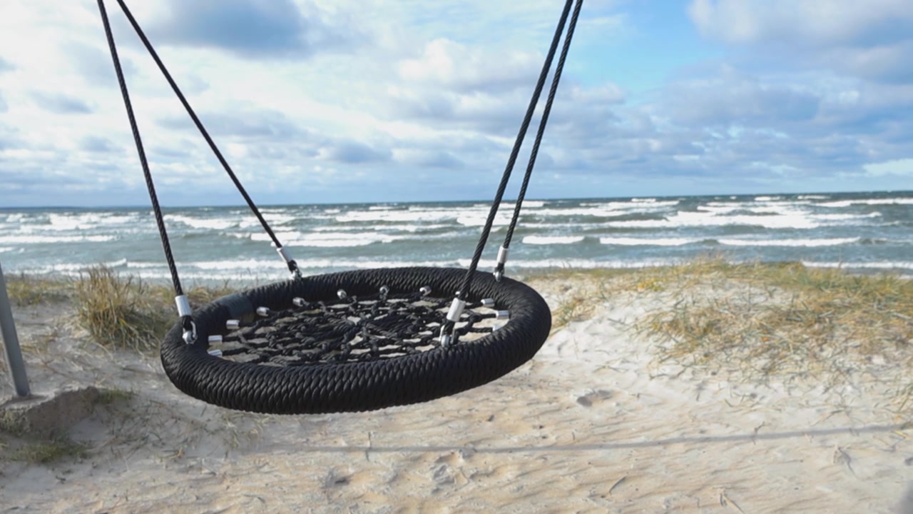 Empty round threaded swing at a beach swinging back and forth in slow motion while ocean sea water waves are coming to the shoreline with white water and wave water splatter. Sandy beach with plants.