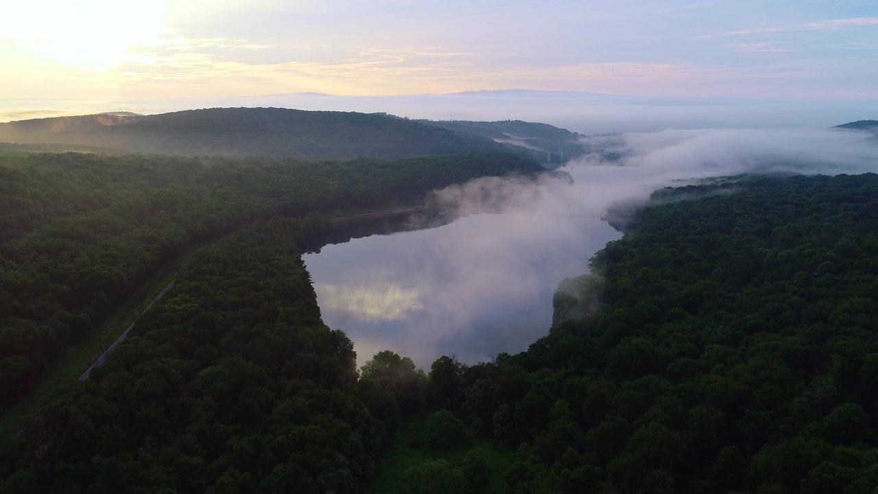 Drone flying forward over a lake and fores during the early morning. A layer of mist-clouds is hovering over the lake