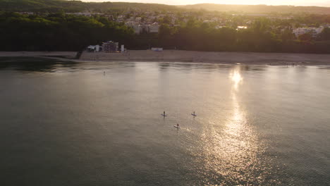 Vista-Aérea-De-Stand-Up-Paddler-En-El-Mar-Báltico-En-Gdynia-Durante-La-Puesta-De-Sol-Dorada,-Polonia---Paisaje-Urbano-En-Segundo-Plano