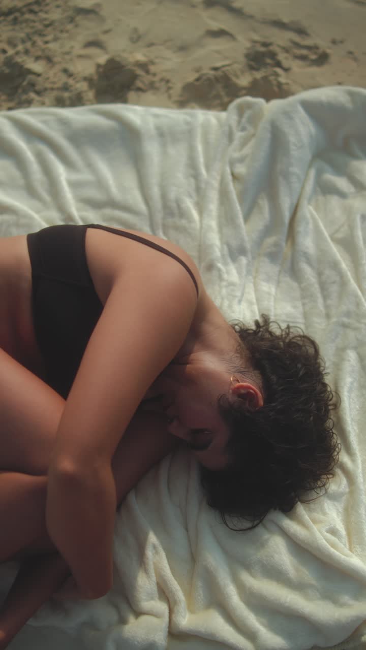 Woman in black bikini lies curled on a blanket, resting peacefully on a sandy beach