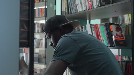 side view of young man in casual clothing wearing cap leaning toward bookshelf, holding book and attentively looking at cover in bright library surrounded by colorful books