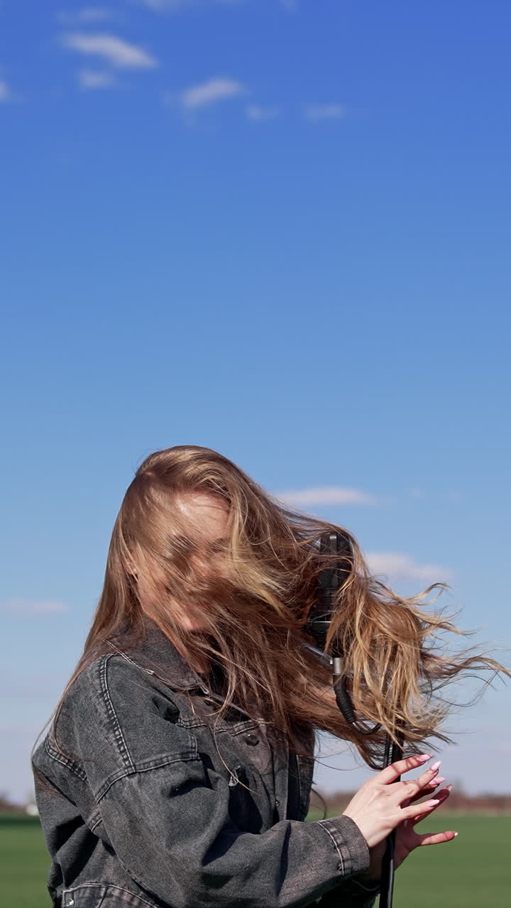 Singer moving her head near the microphone in nature. Attractive girl singing and dancing on green field under blue sky. Vertical video