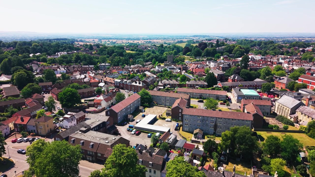 Szeroki widok z lotu ptaka na Devizes w Wiltshire, ukazujący budynki mieszkalne, zabytkowe dachy, wieżę kościelną i zieloną okolicę rozciągającą się poza miasteczkiem targowym w jasny letni dzień.