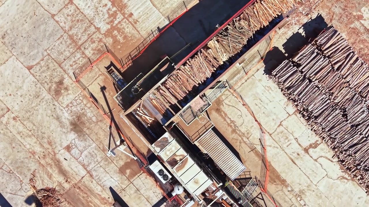Wood processing facility showing logs being handled and prepared for production at a location with concrete surfaces and machinery during daylight hours