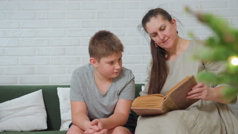 boy in grey top and woman in brown gown sitting on green couch reading old book in cozy room white brick wall background christmas tree partially visible peaceful bonding indoor family moment