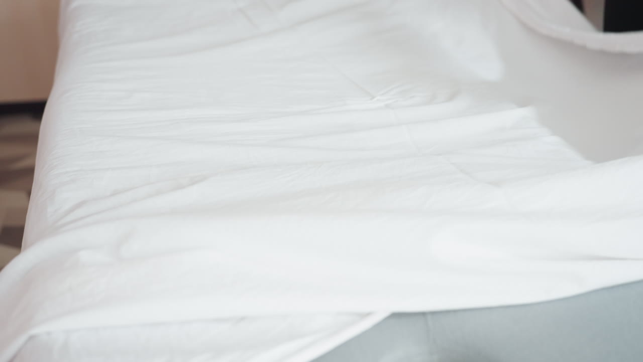 Light skin woman spreads white bed sheet over soft gray mattress in clean interior, motion captured mid-action with fabric lifted in air, highlighting texture, simplicity, and neat bedroom environment