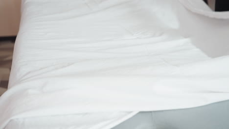 light skin woman spreads white bed sheet over soft gray mattress in clean interior, motion captured mid-action with fabric lifted in air, highlighting texture, simplicity, and neat bedroom environment