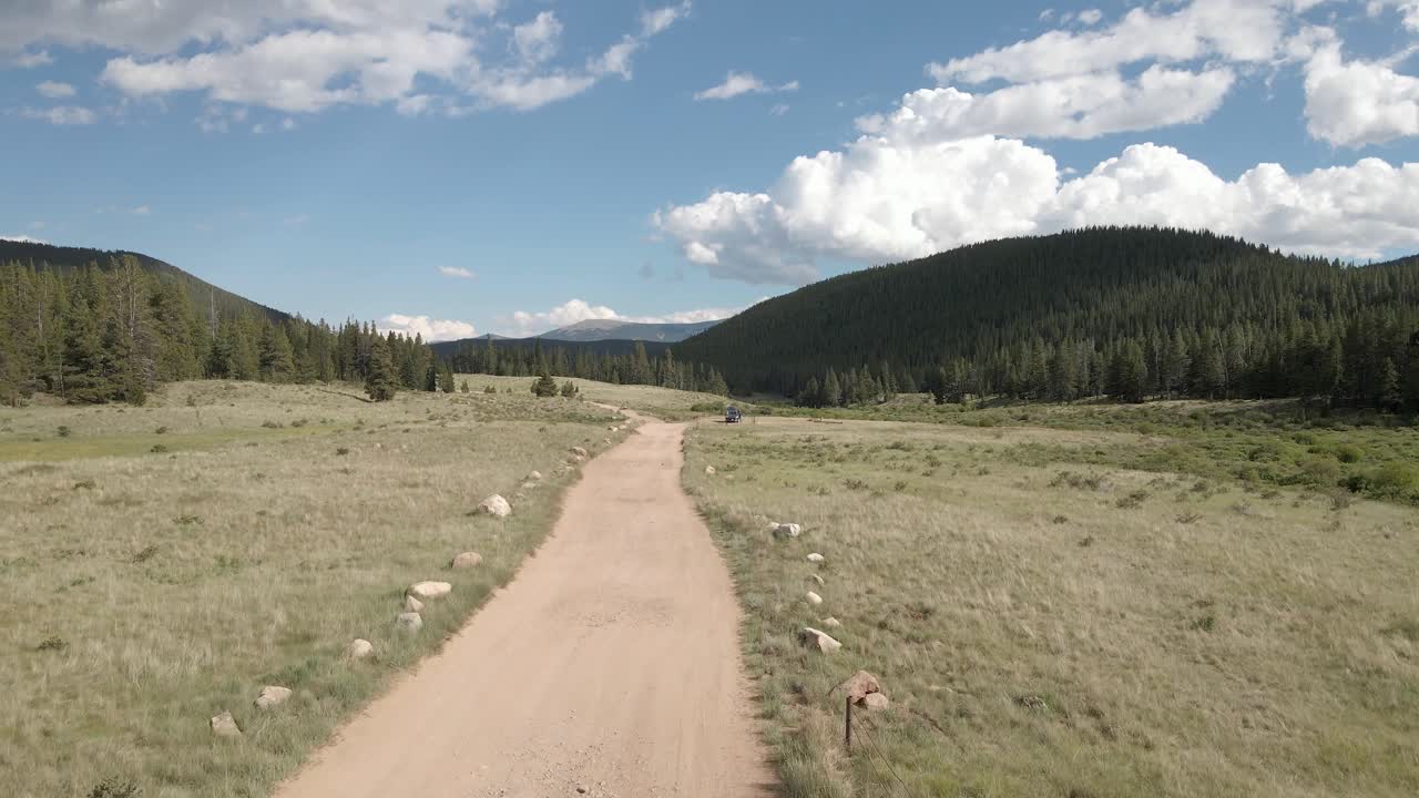 Aerial view following a remote dirt road near Guanella Pass, Colorado. Dolly movement with green grass, forest and clouds in the distance.