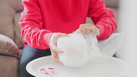 young man sitting on sofa unboxing postal package wrapped in bubblewrap plastic at home