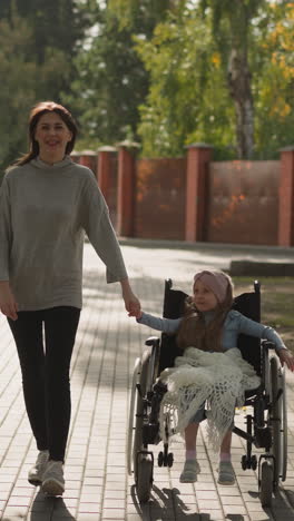 happy woman and little girl join hands enjoying walk in city park on sunny spring day. loving mother looks at daughter sitting in wheelchair