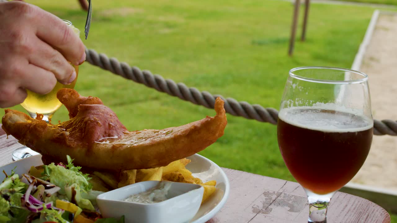 Hand squeezes lemon onto fried fish and chips with beer, natural daylight, outdoor setting