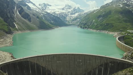 an impressive concrete dam stretches across a vivid turquoise lake, surrounded by majestic snow-covered mountains and green alpine slopes.