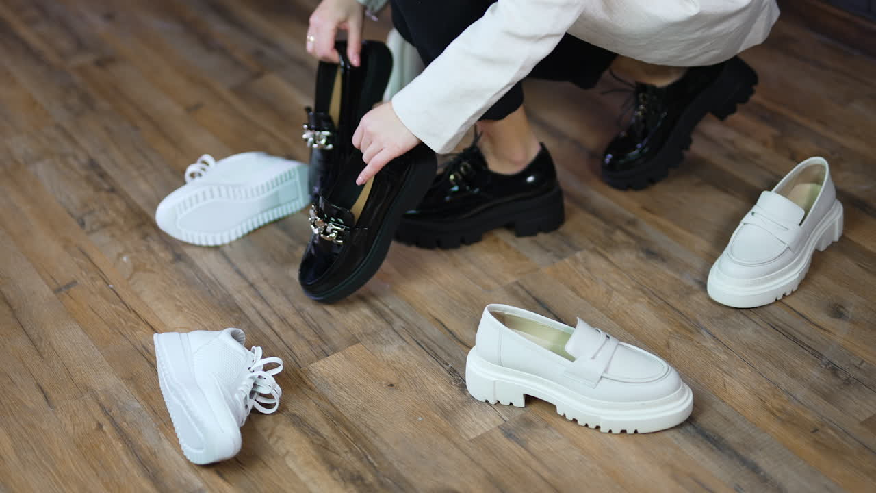 Woman in massive black shoes walks among the scattered footwear. Lady picks up another pair of black shoes leaving white ones on the floor.