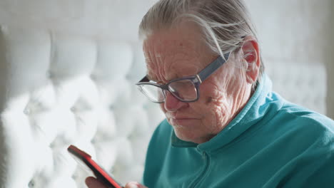 close-up of elderly woman wearing glasses and turquoise fleece sweater, intensely focused on her smartphone. bright natural light from window creates soft background lighting