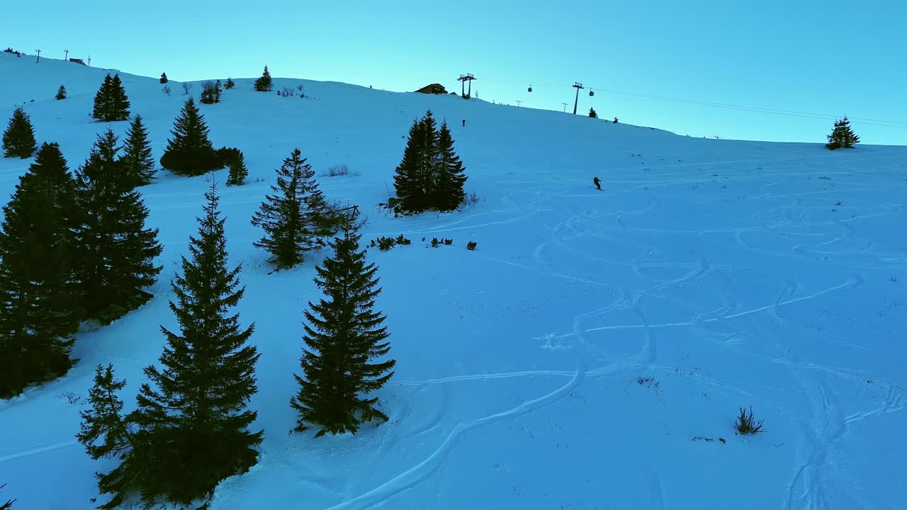 Filmato aereo da drone che si sposta verso destra, riprendendo uno sciatore freestyle che scende lungo un pendio innevato con tracce di sci, alberi sempreverdi e impianti di risalita sotto un cielo invernale azzurro. Posizione: Mala Fatra, Slovacchia