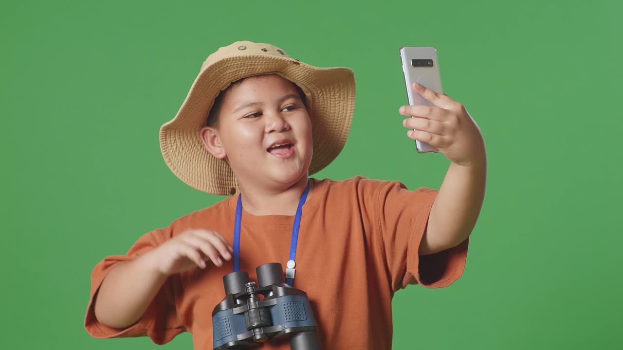 모자와 안경을 들고 손을 흔들며 녹색 스크린 배경에 서서 스마트폰에서 비디오 통화를 하는 아시아 관광 소년. 소년 연구자는 무언가를 조사합니다.