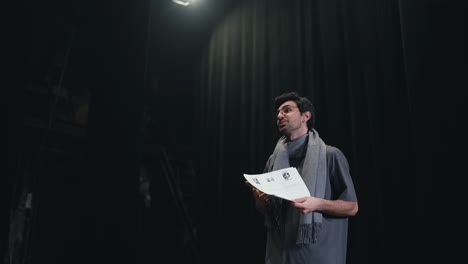 side view of a confident male stage director in a gray t-shirt and scarf walking onto the stage along with a stack of papers and greeting everyone in the theater with black curtains