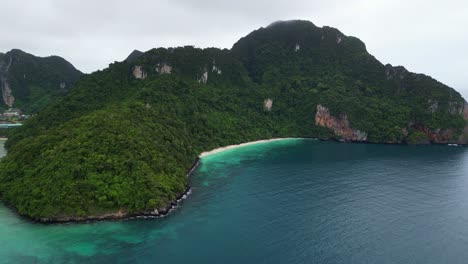 aerial view of lush green hills surrounding a clear blue bay, with a sandy beach visible beneath cloudy skies.