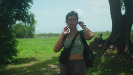 a woman stands in nature, adjusting her hair and glancing sideways. she wears a crop top and carries two bags. the camera captures her in an american shot